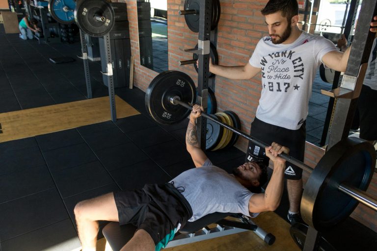 Home A strong man lifts weights on a bench while a trainer assists in a modern gym setup.
